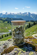 Pot en verre de fleurs de CBD "Green Horizon" posé sur un rocher moussu, surplombant une vallée alpine verdoyante et des montagnes en été.