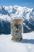 Pot en verre de fleurs de CBD "Mont Blanc OG" posé directement dans la neige, avec le massif du Mont Blanc enneigé et un ciel bleu en arrière-plan.