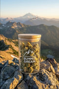 Pot en verre de fleurs de CBD "Summit Dream" posé sur des rochers en haute montagne, avec une vue panoramique sur les sommets au coucher du soleil.